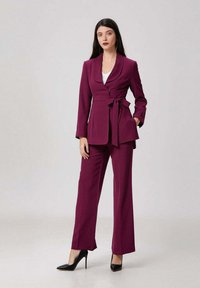 Femme portant un tailleur prune ajusté avec un blazer ceinturé, un haut blanc et des chaussures à talons hauts noires, se tenant contre un fond uni.