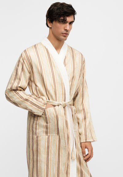Jeune homme portant un peignoir rayé beige et marron avec ceinture nouée, debout, une main dans la poche, sur un fond uni.