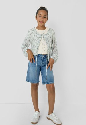 Dziewczyna stojąca z rękami przy kieszeniach jeansowych szortów, ubrana w kwiecistą prześwitującą bluzkę, biały top i białe tenisówki na gładkim tle.