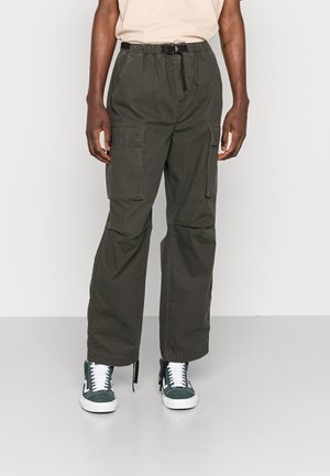 Henkilö, jolla on oliivinvihreät, suurilla sivutaskuilla varustetut cargo-housut, musta säädettävä vyö, beige t-paita ja vihreät tennarit valkoisilla nauhoilla.