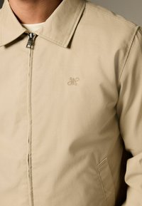 Veste beige avec un design à col, dotée d'une fermeture éclair, de poches latérales et d'un logo brodé discret sur la poitrine. Texture lisse.
