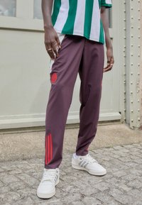 Bordeaux sportbroeken met rode accenten en drie rode strepen aan de zijkanten, gecombineerd met witte schoenen met grijze strepen.