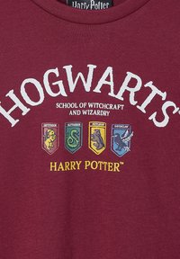 Bordeauxrotes Baumwoll-T-Shirt mit dem Aufdruck "HOGWARTS" in Weiß, geschmückt mit bunten Hauswappen: Gryffindor, Slytherin, Hufflepuff, Ravenclaw.