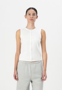 Sleeveless white ribbed top with a crew neckline and button-down front. Worn with gray relaxed-fit sweatpants. Smooth texture and slim fit.