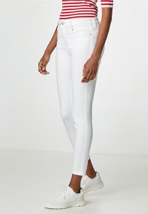 Osoba ubrana w biały, dopasowany dżins, czerwono-białą koszulkę w paski oraz białe trampki, stojąca na tle jednolitego, jasnego tła.