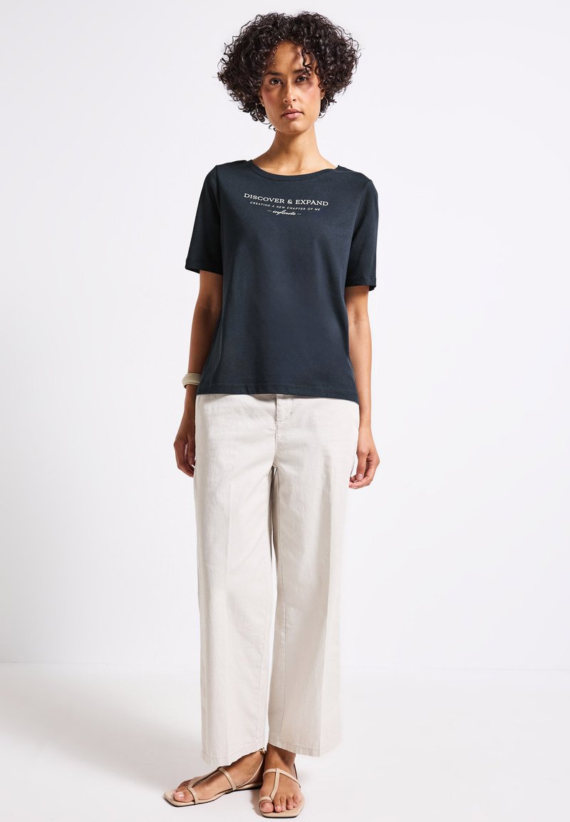 T-shirt noir à manches courtes avec le texte "DÉCOUVRIR & ÉLARGIR" ; porté avec un pantalon large crème ; le modèle se tient sur un fond clair.