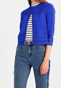 Kvinna som bär en kungligt blå kortärmad cardigan med perforerade detaljer över en svart-vitrandig polotröja och högmidjade blå jeans.