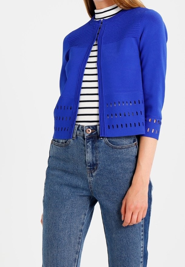Kvinna som bär en kungligt blå kortärmad cardigan med perforerade detaljer över en svart-vitrandig polotröja och högmidjade blå jeans.