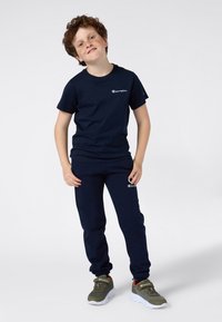 Conjunto de camiseta de algodón azul marino y pantalones de chándal con detalles de logo bordados. El modelo lleva zapatillas verde oliva con suelas blancas.