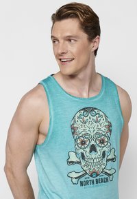 Turkos linne med en färgglad dödskallegrafik och blommönster samt korsade ben; tillverkat av lätt och ventilerande material.