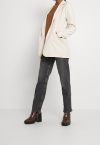 Blazer de color beige claro con solapas de muesca, combinado con una blusa marrón y vaqueros oscuros gris. Botines de plataforma marrón brillante con un diseño elegante.