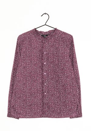 Blouse violette à manches longues avec boutons, motif tacheté blanc et noir, suspendue sur un cintre noir sur fond blanc.