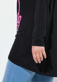 Sweater nero con dettagli grafici rosa sulle maniche, caratterizzato da un fit loose e maniche lunghe. Indossato con jeans blu chiaro. Tessuto morbido.