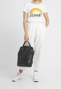 Vrouw in witte high-waist broek, witte sneakers, witte t-shirt met zonsondergangafbeelding, die een zwarte leren handtas met handvatten vasthoudt.