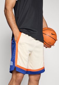 Bēšas basketbola šorti ar ziliem un oranžiem akcentiem, ar svītrainiem sānu elementiem; valkāti ar melnu bezpiedurkņu krekliņu un turēti basketbola bumba.