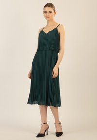 Dunkelgrünes, plissiertes Kleid mit verstellbaren dünnen Trägern, einem V-Ausschnitt und mehrlagigem Design, das bis zur Mitte der Waden reicht. Kombiniert mit schwarzen High Heels.