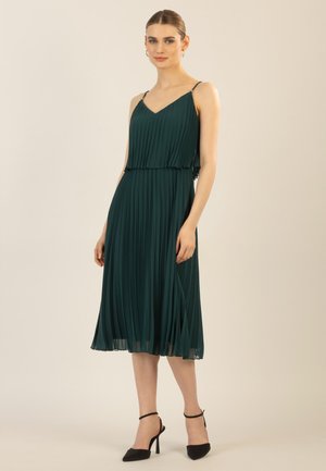 Dunkelgrünes, plissiertes Kleid mit verstellbaren dünnen Trägern, einem V-Ausschnitt und mehrlagigem Design, das bis zur Mitte der Waden reicht. Kombiniert mit schwarzen High Heels.