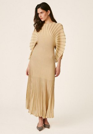 Mujer de pie con un vestido midi de punto acanalado beige con mangas acampanadas y zapatos planos puntiagudos con estampado sobre un fondo liso.