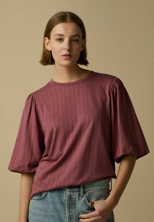 Jeune femme aux cheveux bruns courts portant un haut mauve à manches bouffantes et un jean bleu clair sur un fond beige.