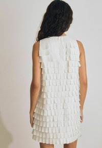 Valge ilma varrukateta kleit, millel on kihiline disain ruudukujuliste volangidega, tekstureeritud kangas ja tagumine lukuga sulgur.