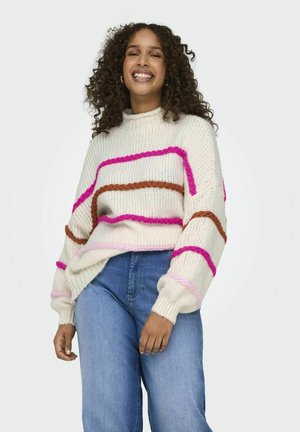 Gebreide trui in crème met dikke roze, bruine en lichtroze strepen; heeft een hoge nek, een losse pasvorm en geribbelde manchetten. Gecombineerd met blauwe jeans.