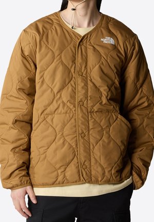 Person iført en brun quiltet jakke med trykknapper og The North Face-logo på brystet, kombineret med sorte bukser og en beige skjorte.