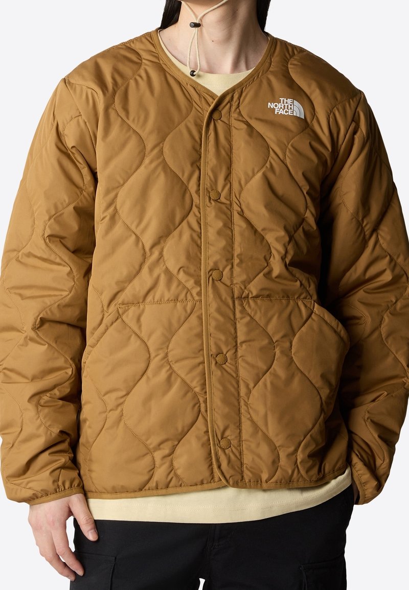 Personne portant une veste marron matelassée avec des boutons-pression et le logo The North Face sur la poitrine, associée à un pantalon noir et une chemise beige.