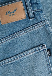 Hellblaue Jeans aus Denim mit einem braunen Lederetikett mit der Aufschrift "Reell" und sichtbaren Nähten in kontrastierendem Orange.