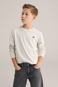 Krém színű hosszú ujjú póló kerek nyakkivágással és fekete logóval a mellkason, szürke farmernadrággal párosítva. Egyszerű kialakítás, sima textúra.