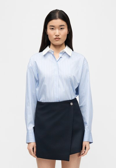 Νεαρή γυναίκα που φοράει ένα ανοιχτό μπλε ριγωτό πουκάμισο με κουμπιά, το οποίο είναι μισοβιδωμένο μέσα σε μια μαύρη ασύμμετρη φούστα τύπου wrap με κουμπί στο πλάι.