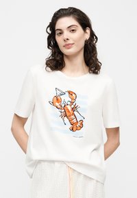 Γυναίκα με σγουρά σκούρα μαλλιά που φοράει λευκό T-shirt με μεγάλο πορτοκαλί γραφικό αστακού πάνω σε ανοιχτό μπλε ρίγες.