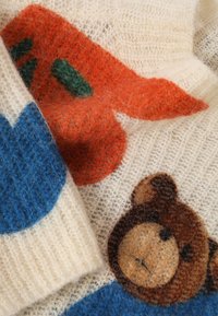 Pleteni pulover v kremni barvi, ki vsebuje pisane vzorce modre, oranžne in zelene barve, z motivom rjavega medveda. Mehak, puhast učinek.