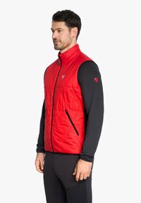 Red insulated vest with diamond stitching, high collar, zippered front, and two side pockets. Worn over a black long-sleeve shirt.
