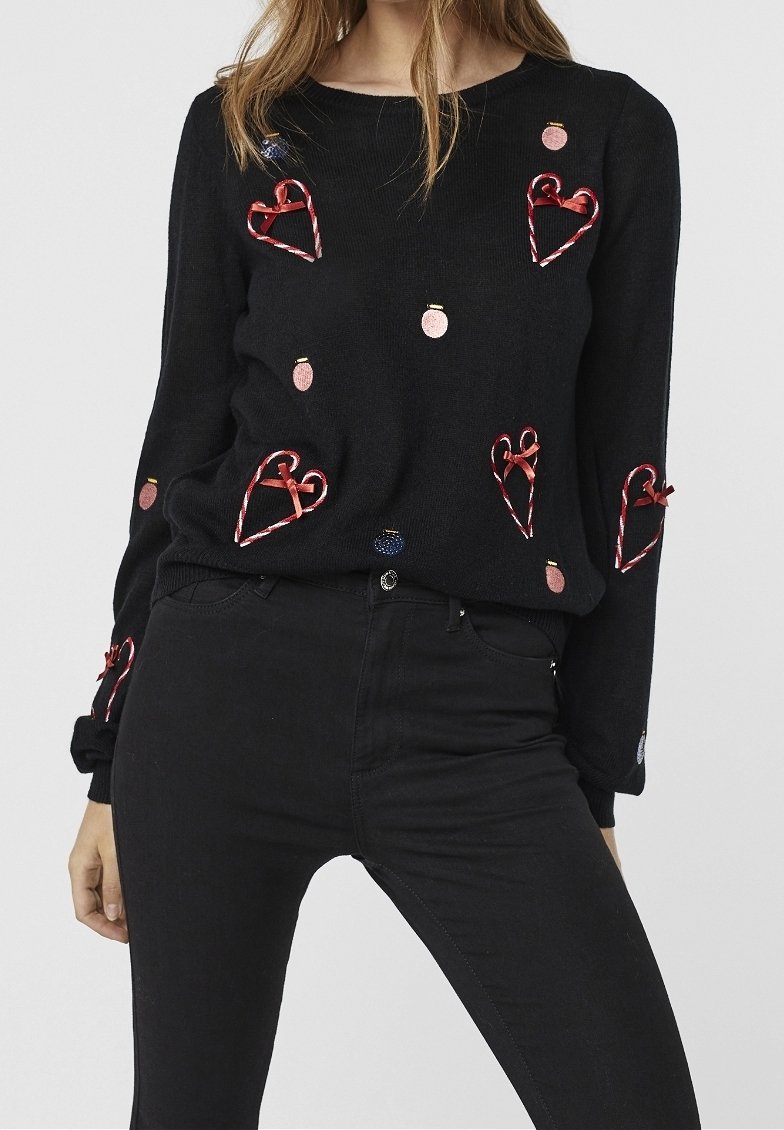 Femme portant un pull noir orné de cœurs en ruban rouge et de petits ornements circulaires roses et bleus, assorti à un pantalon noir.