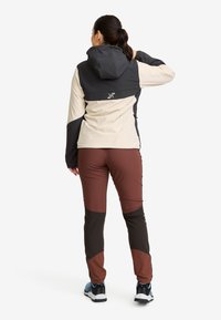 Woman standing with back facing, wearing a black and beige hooded jacket, brown and black pants, and blue sneakers on a white background.