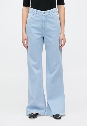 Φαρδιά τζιν παντελόνια με ψηλή μέση σε ανοιχτό μπλε χρώμα, φορεμένα με μαύρο τοπ και μαύρα παπούτσια με ασημί οπές, εμφανιζόμενα από τη μέση μέχρι τα πόδια.