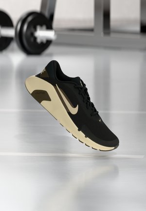 Czarno-beżowy but do biegania Nike zawieszony w powietrzu z rozmytym tłem siłowni i ciężarami.