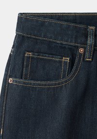 Poche de jean en denim bleu foncé avec surpiqûres orange, rivets métalliques et passants de ceinture sur la ceinture, sur un fond clair.