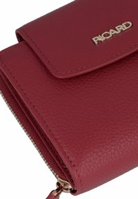 Cartera de cuero rojo con acabado texturizado, logo dorado, cierre de cremallera y bordes cosidos. Diseño compacto con solapa superpuesta.