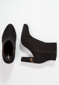 Bottines en daim noir avec talons carrés, fermetures éclair latérales et petite broderie florale marron sur le talon, vues de dessus et de côté.