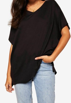 Kobieta w luźnej czarnej koszulce z dekoltem w serek i jasnoniebieskich dżinsach, z jedną ręką w kieszeni oraz złotym naszyjnikiem z okrągłym wisiorkiem.