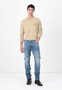 Beiger Strickpullover mit kleinem rotem Logo, kombiniert mit hellblauen Denim-Jeans. Das Modell trägt schwarze Schuhe mit einer glatten Oberfläche.