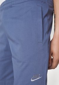 Nike tepláky v tlumené modré barvě, s boční kapsou, elastickým pasem a vyšitým logem ve stříbrné barvě na nohavici.