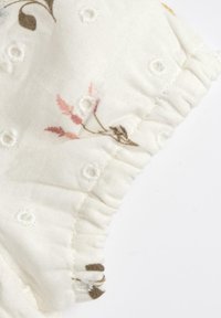 Tissu léger blanc avec un motif floral en rose et marron. Présente des broderies texturées et des détails de volants froncés le long du bord.