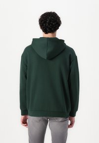 Hoodie vert foncé avec une poche kangourou, des poignets et un ourlet côtelés. Fabriqué en tissu doux avec une capuche dotée d'un cordon de serrage en boucle.