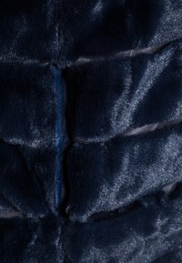 Tissu en fausse fourrure bleu marine avec une texture brillante, présentant des segments cousus pour la structure et un minimum de quincaillerie visible. Vue en gros plan.
