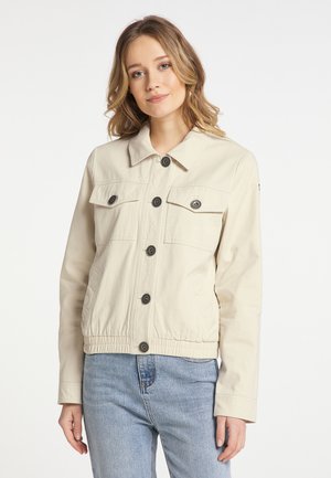 Jeune femme aux cheveux châtain clair portant une veste crème boutonnée et un jean bleu clair sur fond blanc uni.