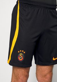 Shorts de sport noirs avec une bande latérale jaune, arborant un logo brodé avec quatre étoiles et un Swoosh Nike orange.