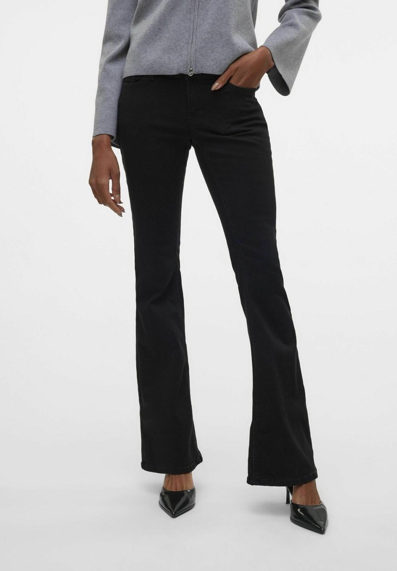 Femme portant un pantalon noir bootcut ajusté, un pull gris à fermeture éclair et des chaussures à talons hauts pointues noires, debout avec une main dans la poche.