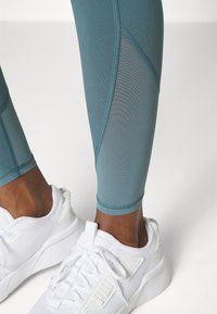 Tealfärgade leggings med en texturerad design och nätpaneler, kombinerat med vita sportskor som har en texturerad sula och logotryck.